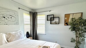 Un dormitorio con cama, un ventanal con persianas, una planta en maceta y obras de arte en las paredes.