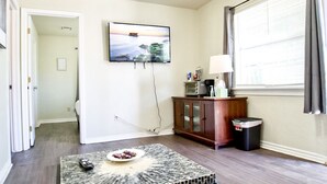 Ein Wohnzimmer mit einem an der Wand befestigten Fernseher, einem kleinen Tisch mit einer Fernbedienung, einem Schrank und einem Fenster mit Vorhängen.