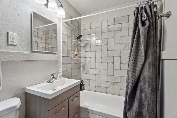 A bathroom with a white sink, wooden vanity, a mirror, a shower with a grey shower curtain, and a tiled wall.