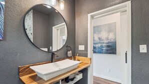 A bathroom with a round mirror, white sink, wooden countertop, and a door with a painting.