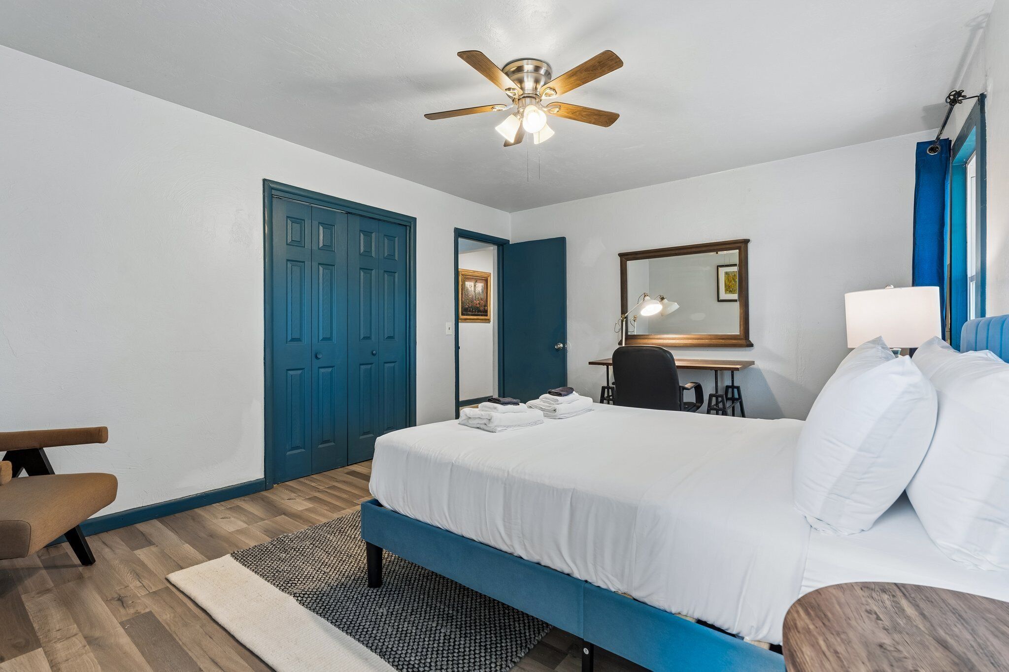 A bedroom with a bed, a desk, a chair, a ceiling fan, a mirror, and a door.