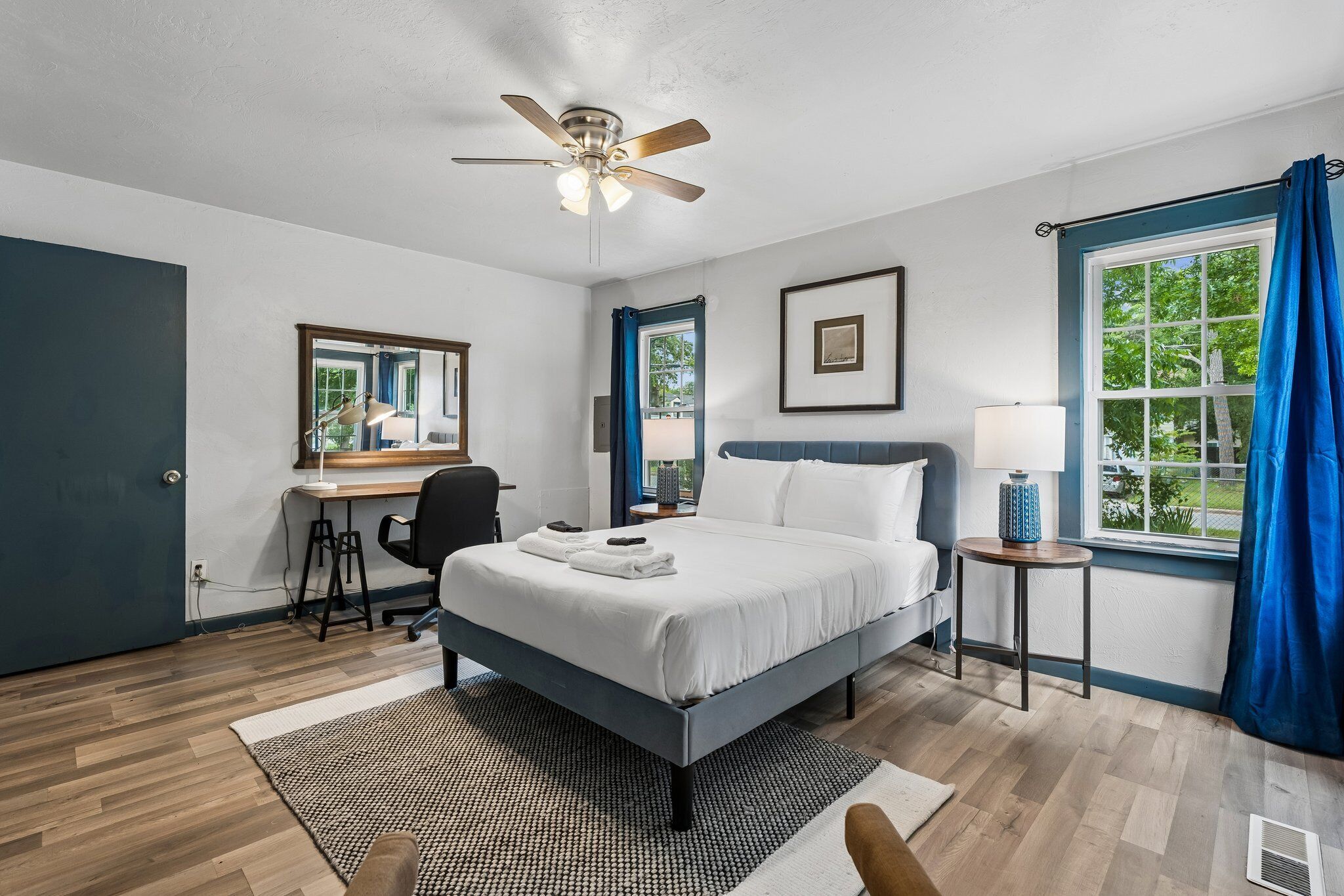 A bedroom with a bed, a desk, a chair, a ceiling fan, a window with curtains, and a view of greenery outside.