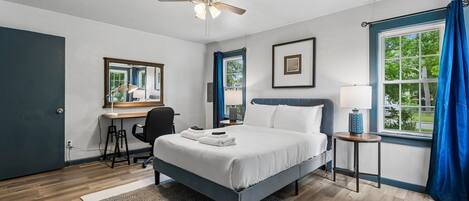 A bedroom with a bed, a desk, a chair, a ceiling fan, a window with curtains, and a view of greenery outside.