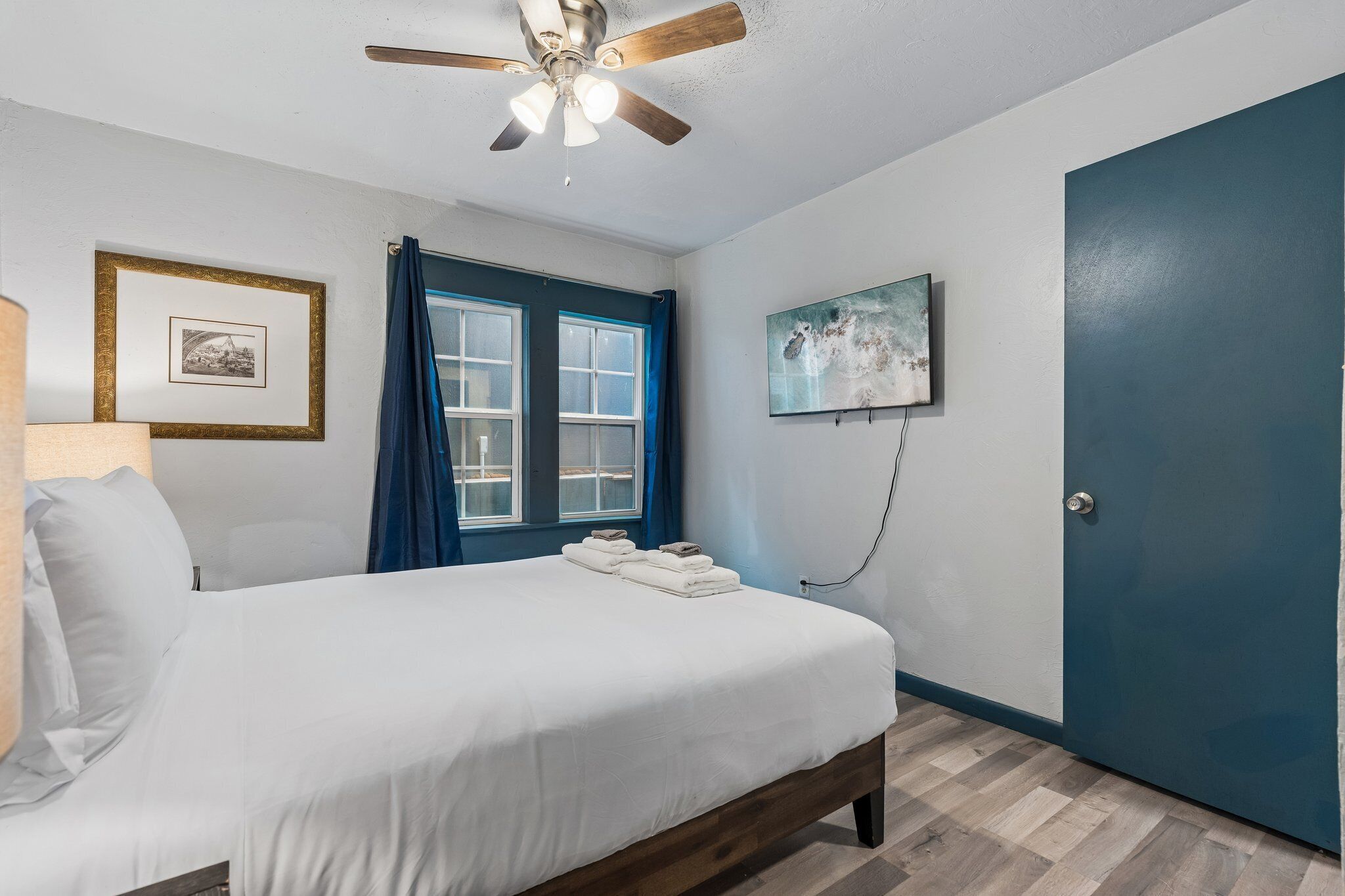 A bedroom with a bed, a ceiling fan, a window with curtains, a door, and a wall-mounted picture.