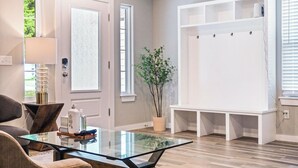 A modern living room with a glass-top coffee table, a gray sofa, a potted plant, and a white entertainment unit with hooks. - 5BR Bethany home, Sleeps 10 — hot tub, full kitchen, near Wiley Post & SNU. (Oklahoma)