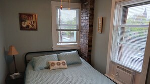 A bedroom with a bed, a nightstand, a lamp, a window with blinds, a brick wall, and a painting on the wall. - Cozy Apartment in the Heart of Downtown Geneva (Geneva)