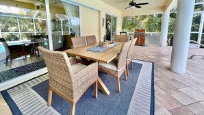 Een terras buiten met een eettafel, rieten stoelen, plafondventilatoren en een barbecue.