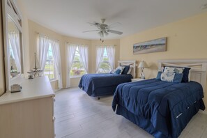 Une chambre avec deux lits, un ventilateur de plafond et un tableau représentant des maisons de plage.