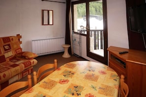A room with a sofa, a table with a floral tablecloth, a television on a wooden stand, and a sliding glass door leading to an outdoor area.