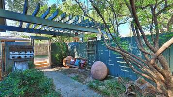 A covered outdoor area with a wooden pergola, a wooden bench with cushions, and a large tree trunk leaning against a blue fence.