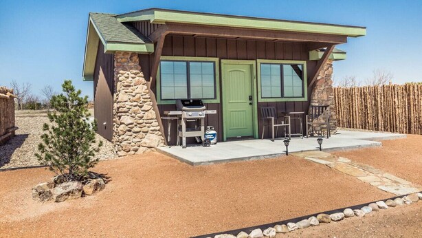 A small, single-story building with a stone exterior and a green door. It features a barbecue grill and two chairs outside.
