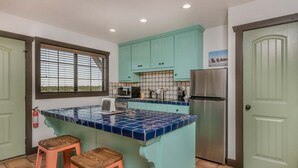 A kitchen with teal cabinets, a blue tiled island, a stainless steel refrigerator, and a window with blinds.