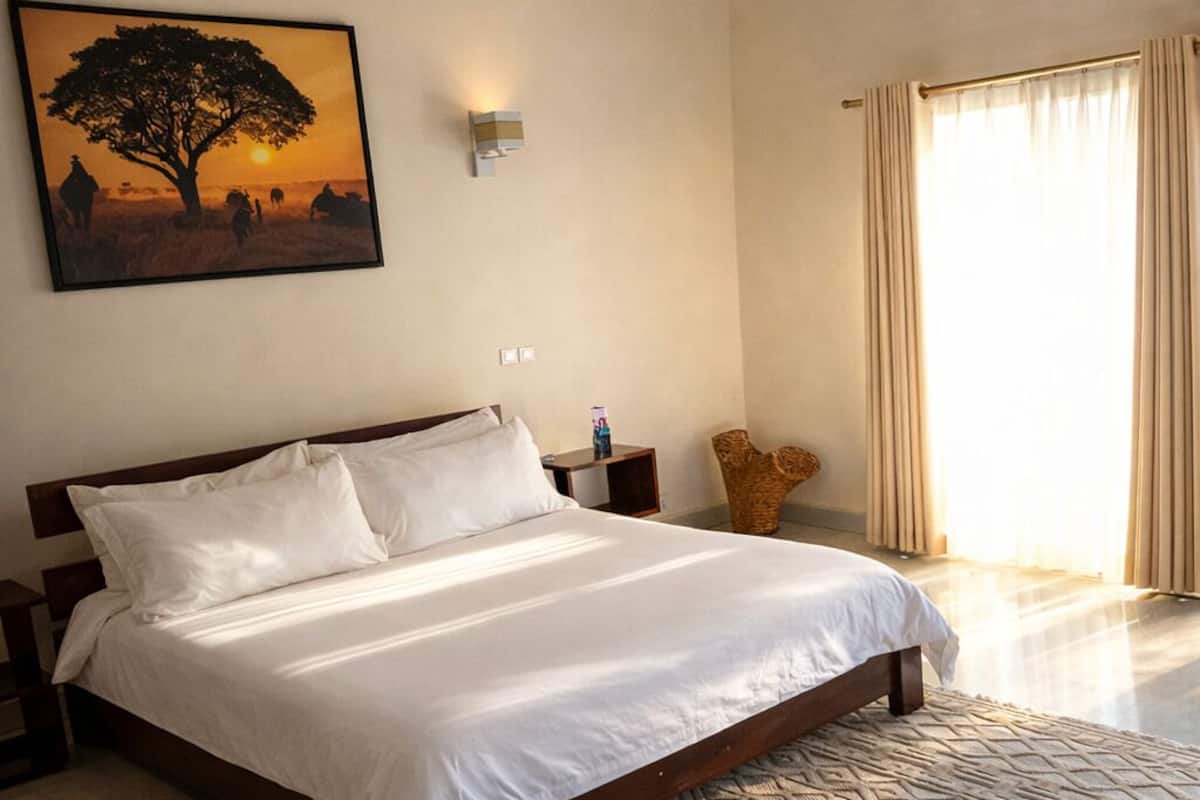 A bedroom with a wooden ceiling, a bed, a nightstand, a wall-mounted light, a painting, and a large window with curtains.