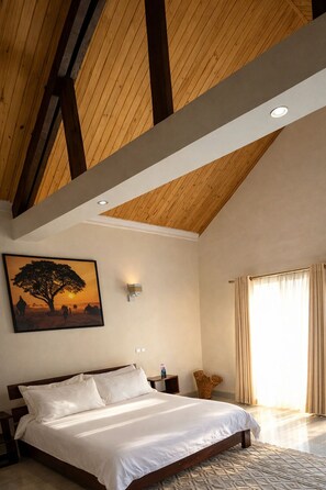 A bedroom with a wooden ceiling, a bed, a nightstand, a wall-mounted light, a painting, and a large window with curtains. - Mahafaly Hotel & Resort (Antsirabe)