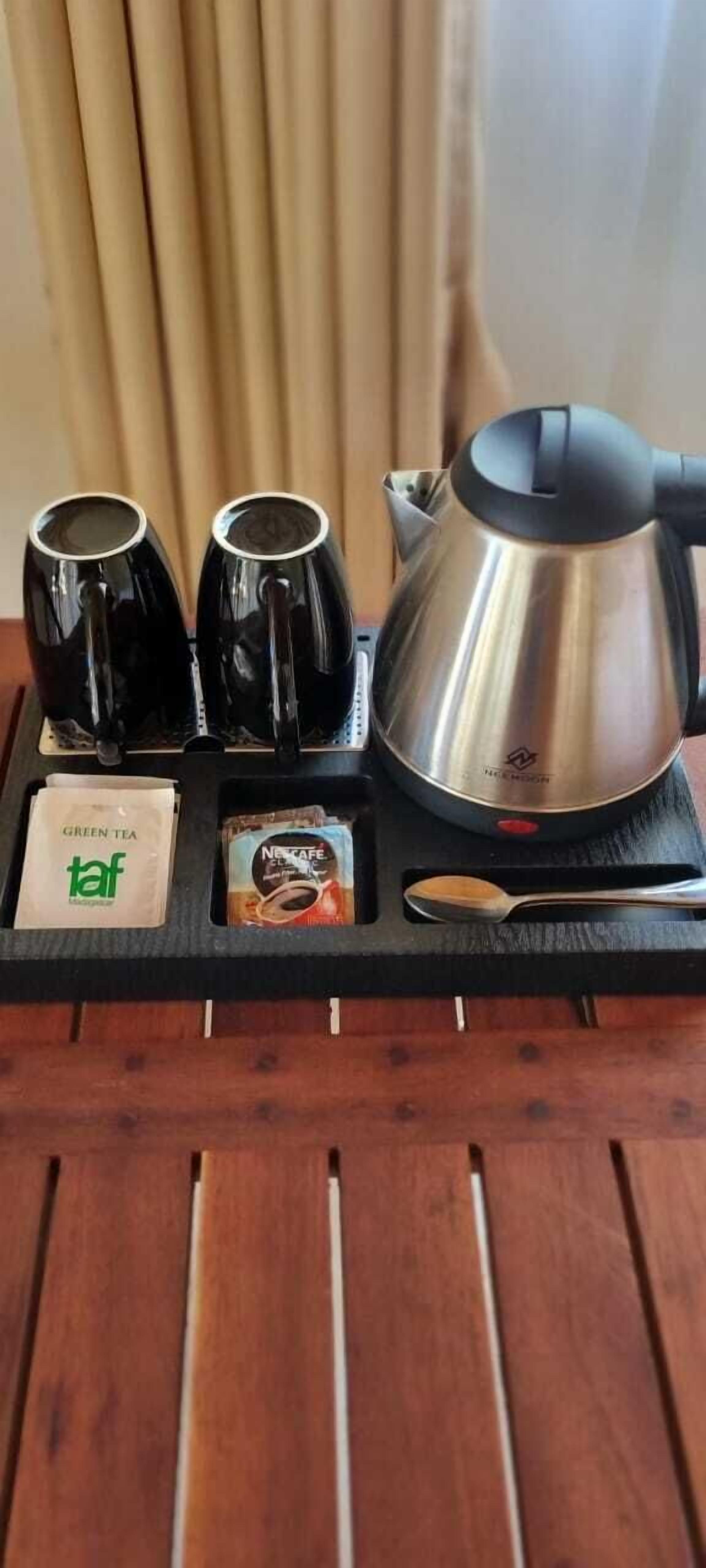 Bandeja con una tetera de acero inoxidable, dos tazas negras, una cuchara y dos bolsitas de té sobre una mesa de madera.