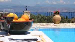 A dining table with a bowl of fruit, a glass of wine, and cutlery, overlooking a pool with a view of mountains. - Villa Bacchus (Corfu)