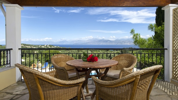 A covered outdoor seating area with wicker chairs and a table, overlooking a scenic landscape with mountains and a body of water. - Villa Bacchus (Corfu)