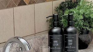 Un lavabo de salle de bain avec un robinet moderne, deux flacons de lotion pour les mains et de savon liquide sur un plateau en bois, et un verre transparent à côté.