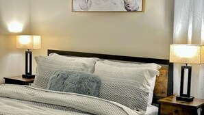 Un lit bien fait, recouvert d’une couverture à motifs, avec deux lampes de chevet et un tableau abstrait accroché au mur.
