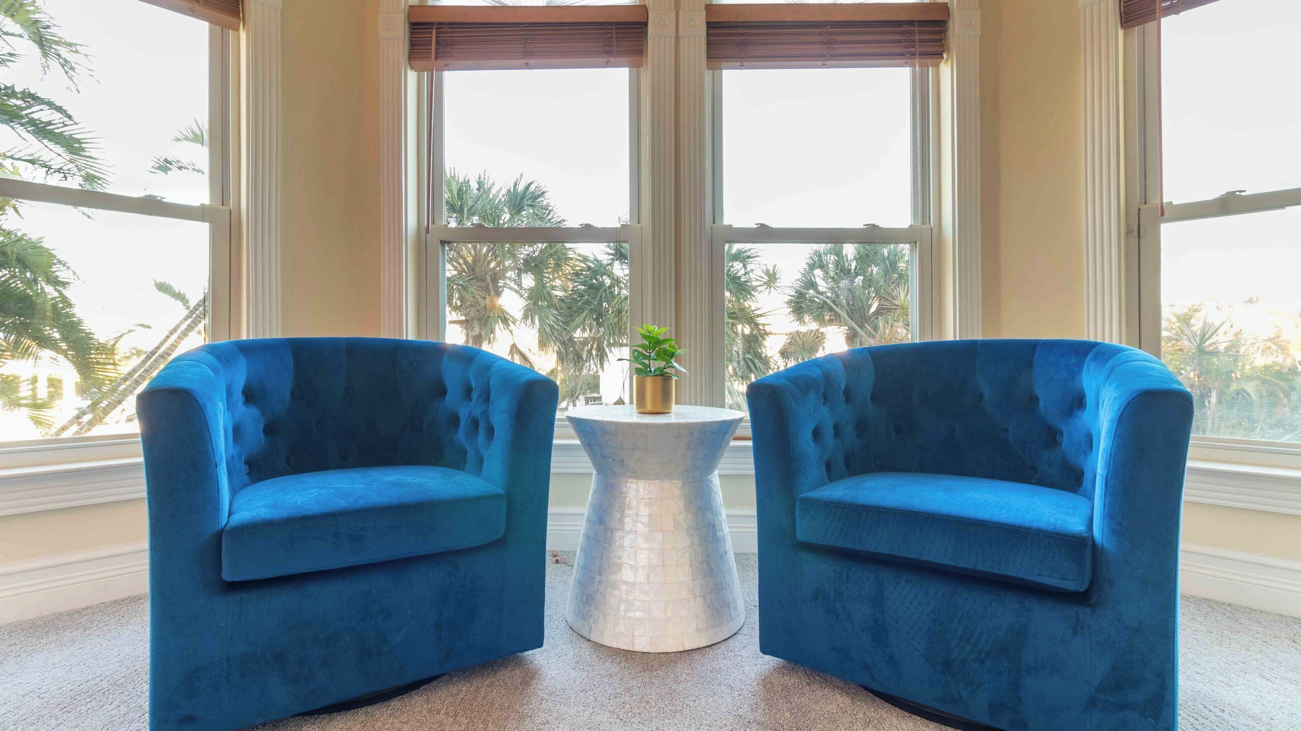 Dos sillones azules enfrentados, con una pequeña mesa redonda entre ellos, ventanas amplias con vista a palmeras y una planta decorativa sobre la mesa.