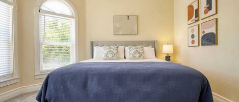 A bedroom with a large bed, a window with blinds, a wall-mounted lamp, and framed artwork.