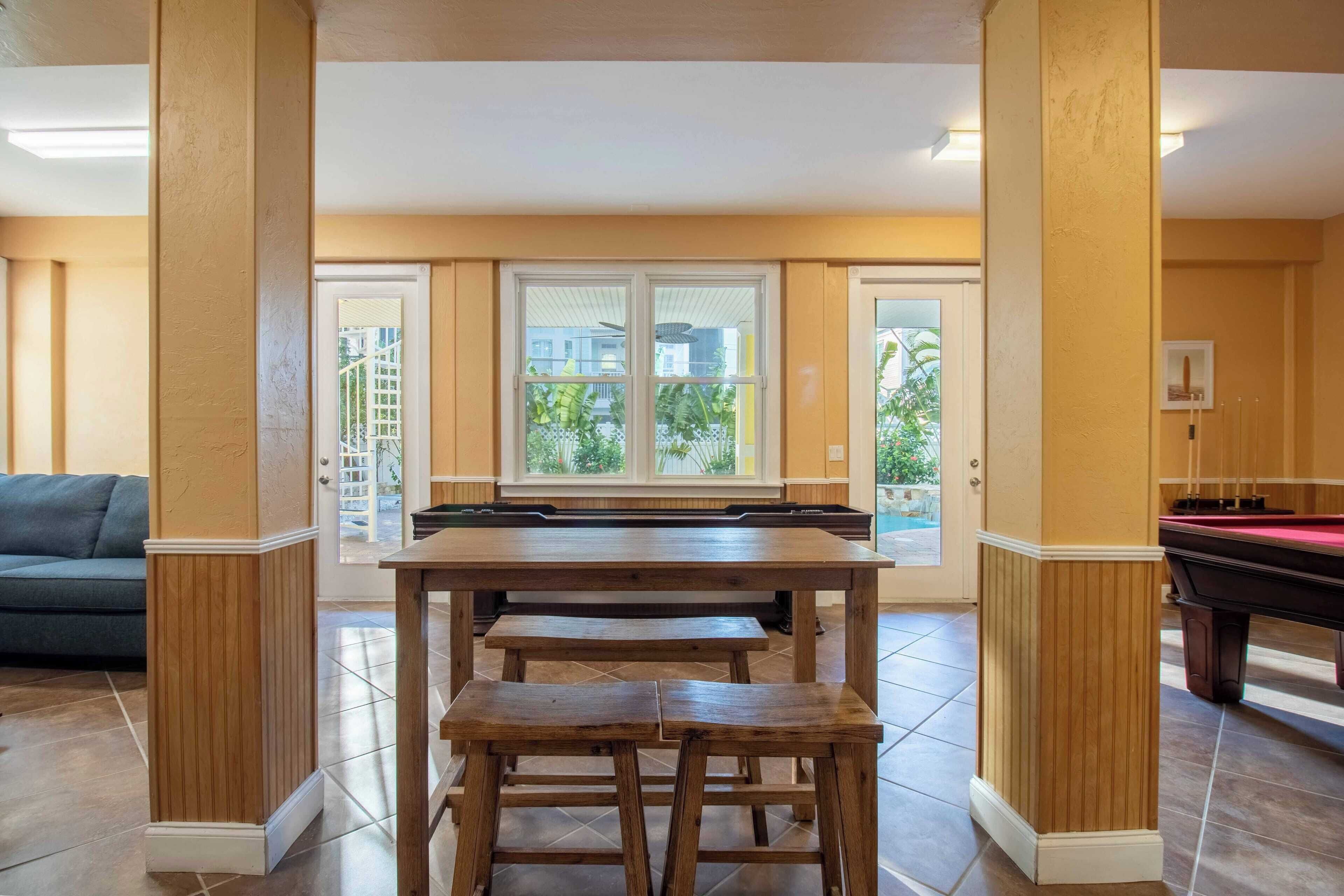 Un espace repas avec une table et des bancs en bois, un billard et une vue à travers une porte vitrée.