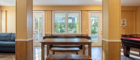 Un espace repas avec une table et des bancs en bois, un billard et une vue à travers une porte vitrée.