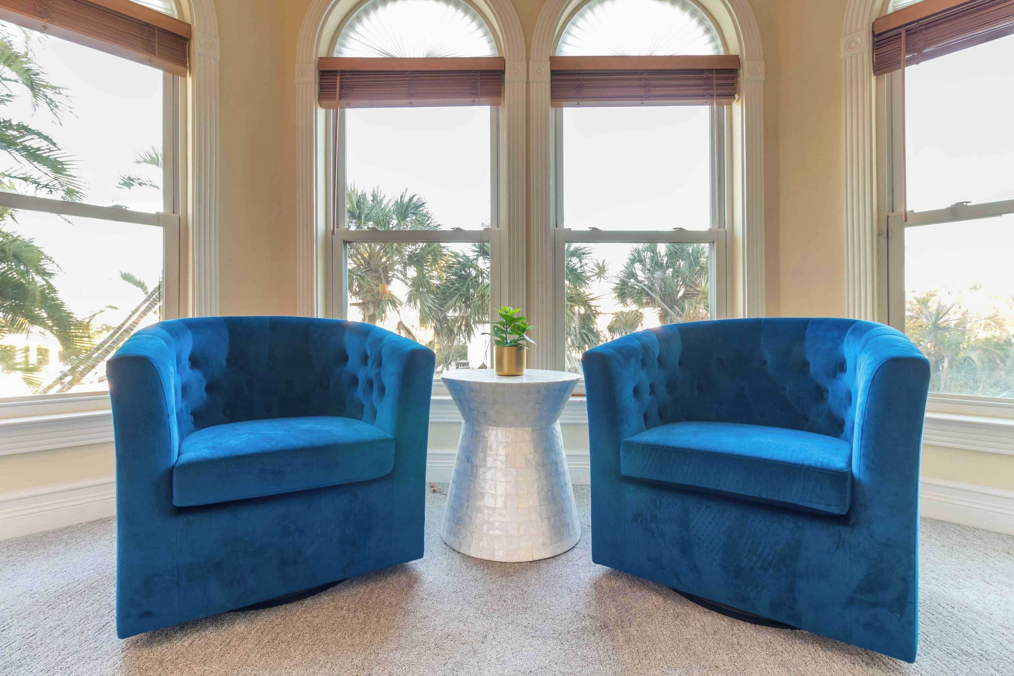 Une pièce avec deux fauteuils bleus placés face à une petite table blanche sur laquelle se trouve une plante. De grandes fenêtres en arc de cercle offrent une vue sur les palmiers à l’extérieur.