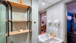 Une salle de bain avec une étagère en bois, une petite plante en pot, un miroir, un lavabo et deux sèche-cheveux.