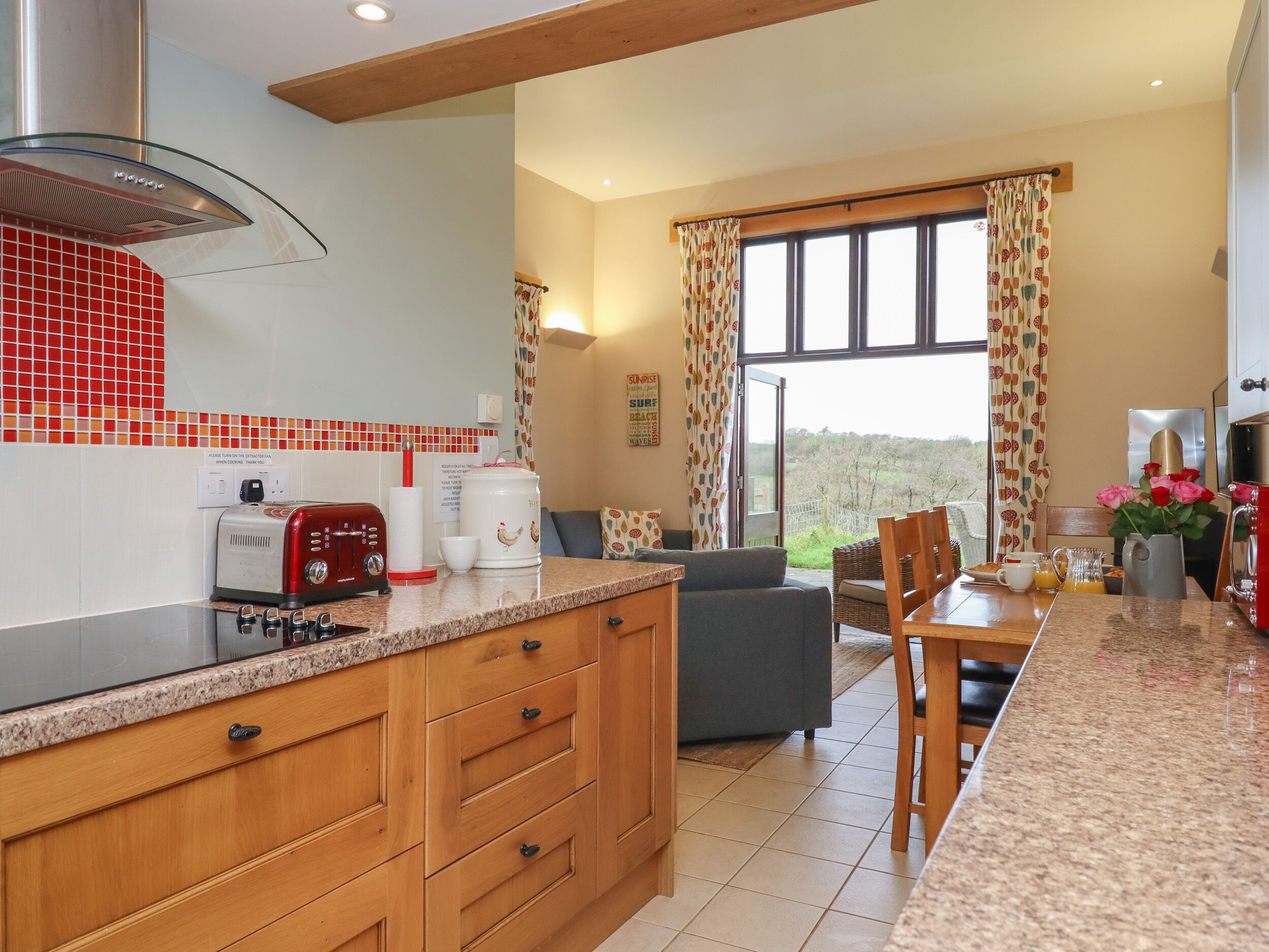 Una cocina moderna con estufa integrada, tostadora y cafetera. Hay un comedor con mesa y sillas, y un ventanal con cortinas.