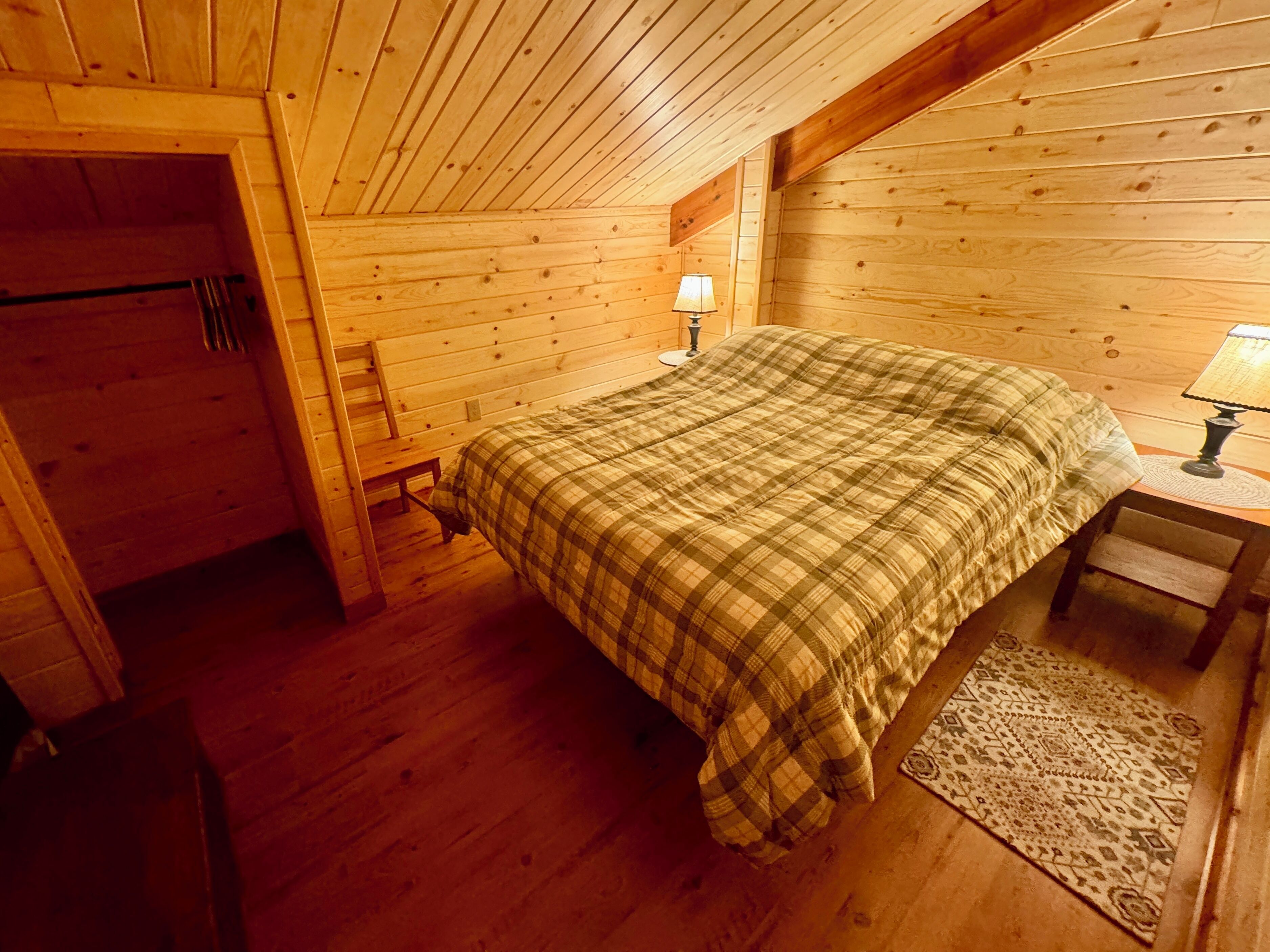 A wooden cabin bedroom with a bed, a nightstand, a lamp, a bench, and a door.