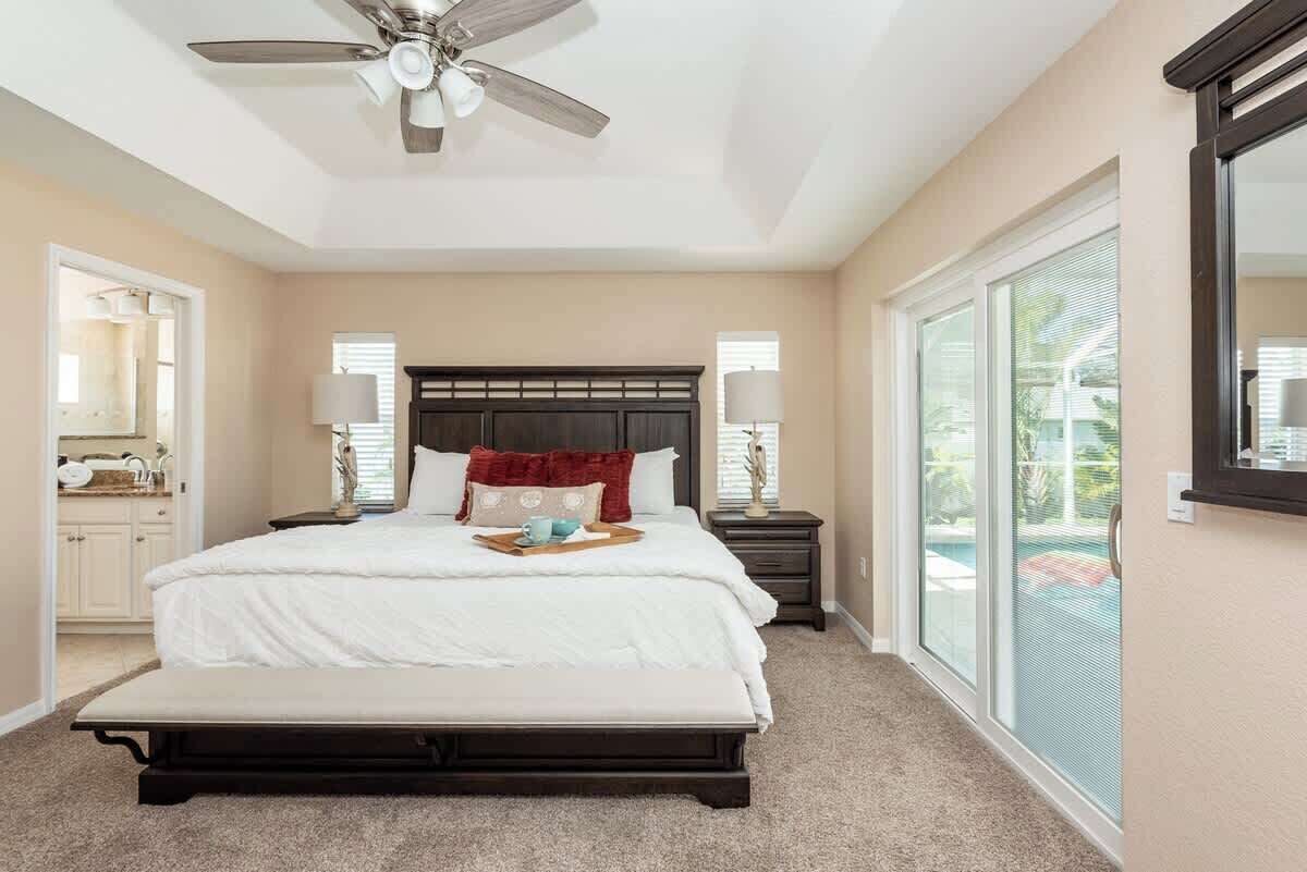 A bedroom with a large bed, a ceiling fan, a nightstand, a lamp, a bench, and a sliding glass door to the outside.