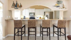 Une cuisine avec des meubles blancs, un four à micro-ondes et une cuisinière. On y trouve trois tabourets de bar en osier, avec des assises tissées.