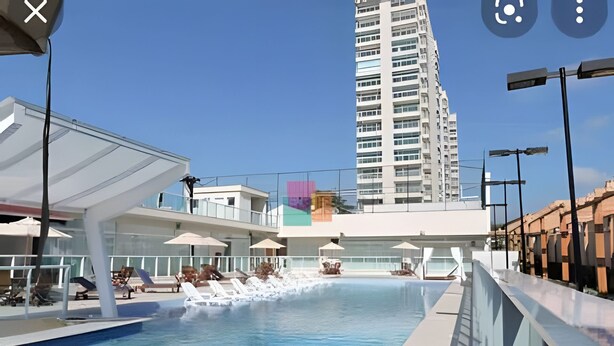 A swimming pool with lounge chairs and umbrellas, surrounded by modern buildings. - Velutti Pérola do Mar - Penha - SC (Penha)