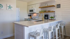A modern kitchen with a central island, white bar stools, a microwave, a coffee maker, and decorative starfish art on the wall. - 30-A Loft with Gulf Views (Santa Rosa Beach)