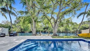 Une piscine rectangulaire à l’eau d’un bleu limpide, entourée d’arbres et bordée d’une terrasse en béton avec des chaises.