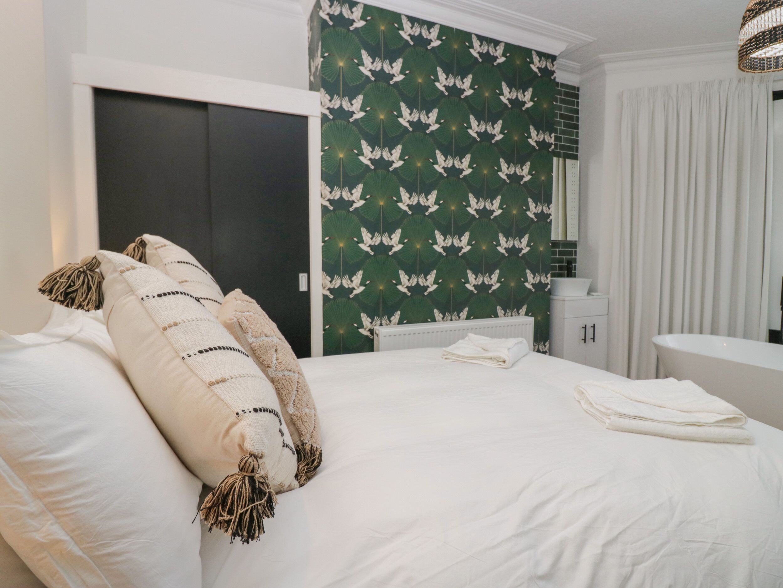 A neatly made bed with white linens and decorative pillows, a dark door, and a green wallpaper with white birds.