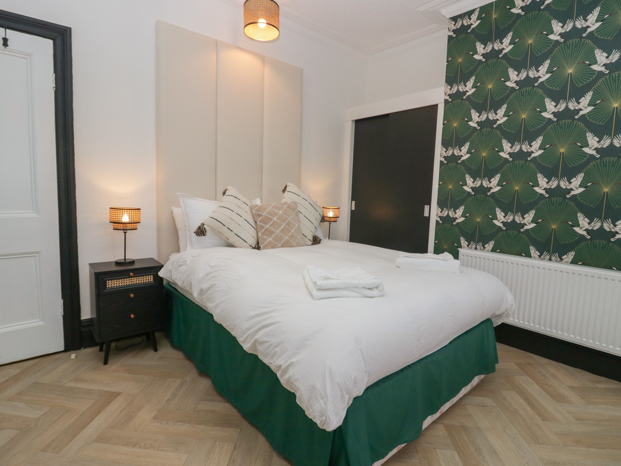 Un lit bien fait, avec une couvre-lit verte, deux tables de chevet avec des lampes, une porte sombre et un mur orné d’un motif d’oiseaux.