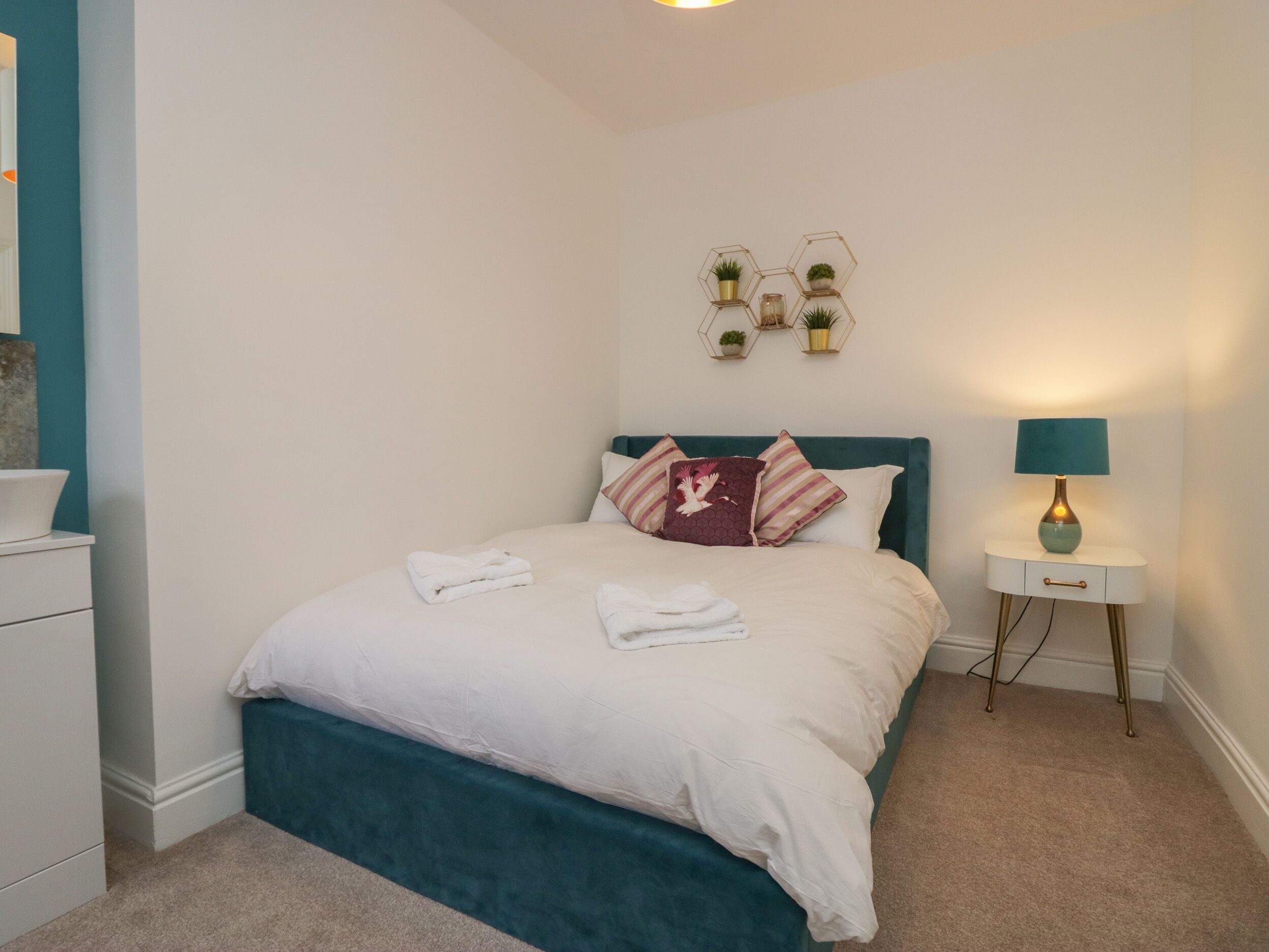 A neatly made bed with white linens and a teal headboard, a bedside table with a lamp, and a wall-mounted plant shelf.