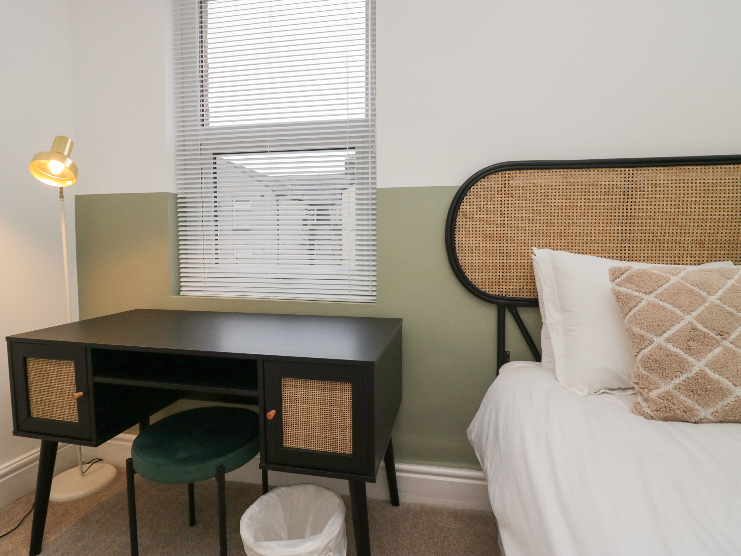 A bedroom with a bed, a desk with a chair, a waste basket, and a window with blinds.