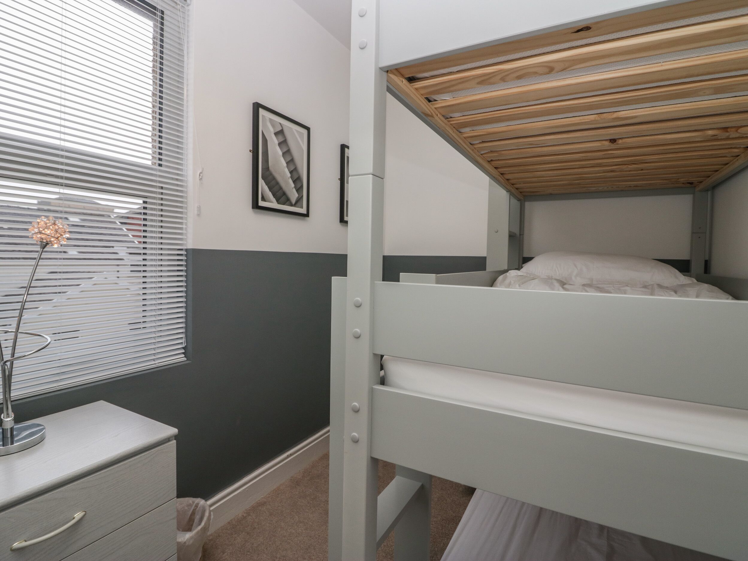 Una cama litera con cabecera de madera, una mesita de noche con cajones, un cuadro enmarcado en la pared y una ventana con persianas.