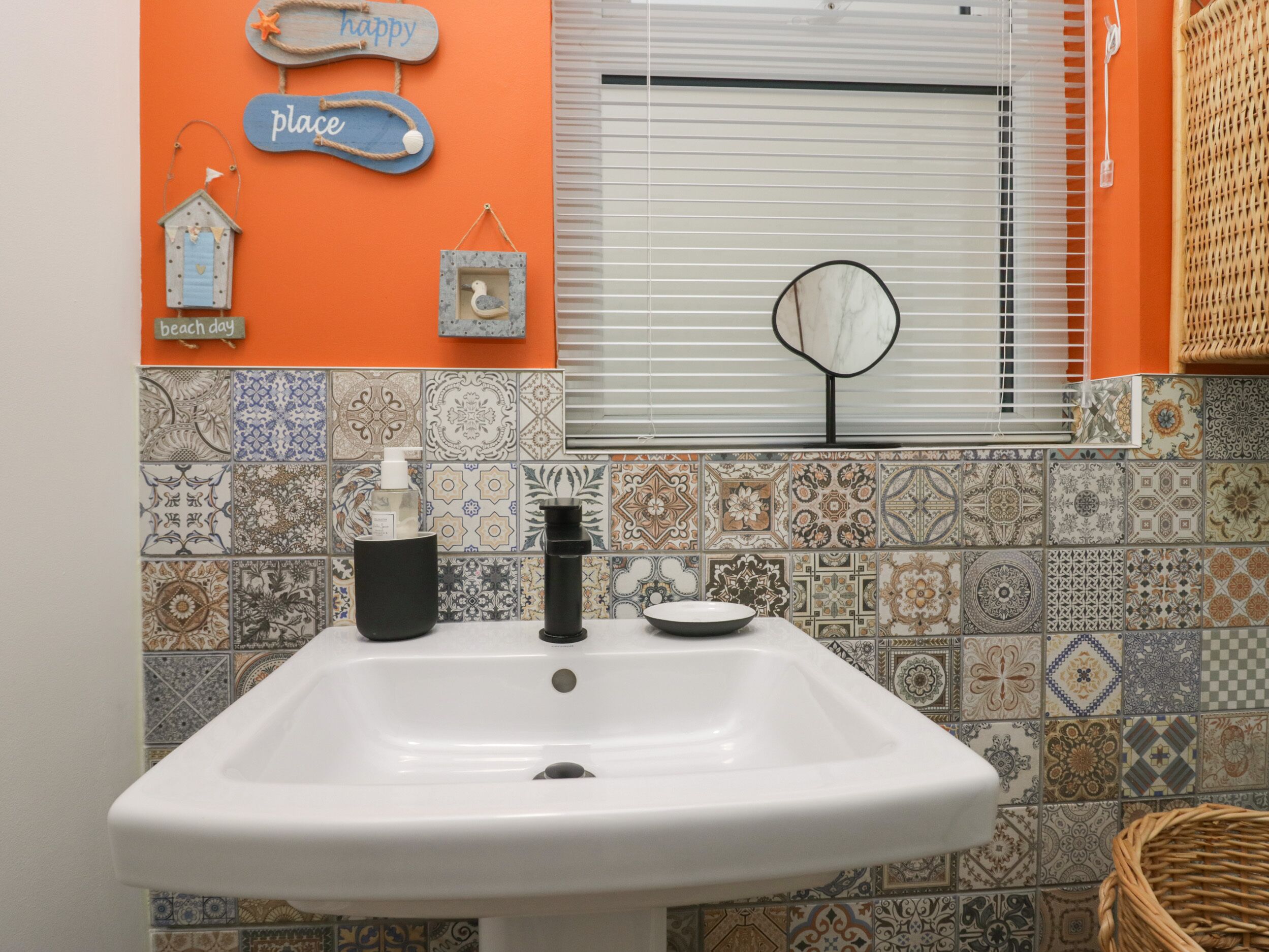 A bathroom with a white sink, decorative tiles, a window with blinds, and wall decorations including a 'happy place' sign and a birdhouse.