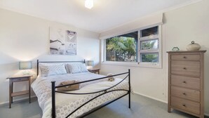 Een slaapkamer met een bed, nachtkastjes, een dressoir, een raam met uitzicht op palmbomen en een wanddecoratie.