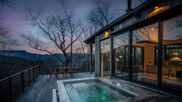 A modern outdoor hot tub area with a wooden deck, glass walls, and a view of the mountains.