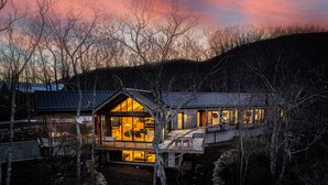 A modern house with large windows and a balcony, surrounded by trees and a rocky landscape.
