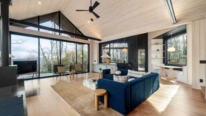 A spacious living room with a large sofa, a coffee table, and a TV. There are large windows offering a view of the outdoors.