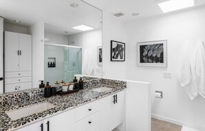 Ein modernes Badezimmer mit einem großen Spiegel, zwei Waschbecken und einer Dusche. Handtücher hängen an der Wand und über dem Spiegel hängt ein gerahmtes Bild.