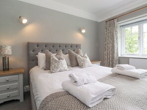 A neatly made bed with a grey headboard, multiple pillows, and a folded white towel on the bed. There is a bedside table with a lamp and a drawer. - Grassings (Ambleside)