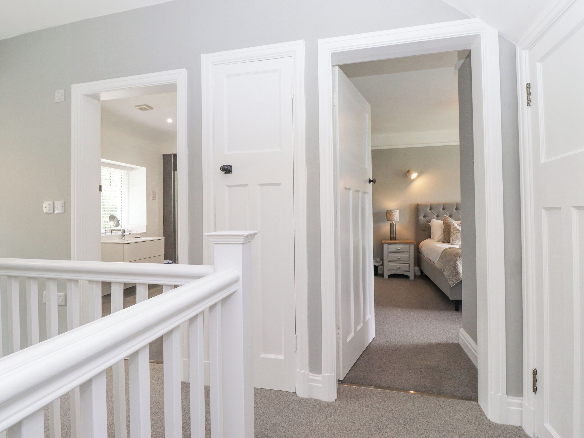 A hallway leading to a bedroom with a bed, bedside table, and lamp. There is a door on the left and a staircase with a white railing.