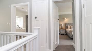 A hallway leading to a bedroom with a bed, bedside table, and lamp. There is a door on the left and a staircase with a white railing.